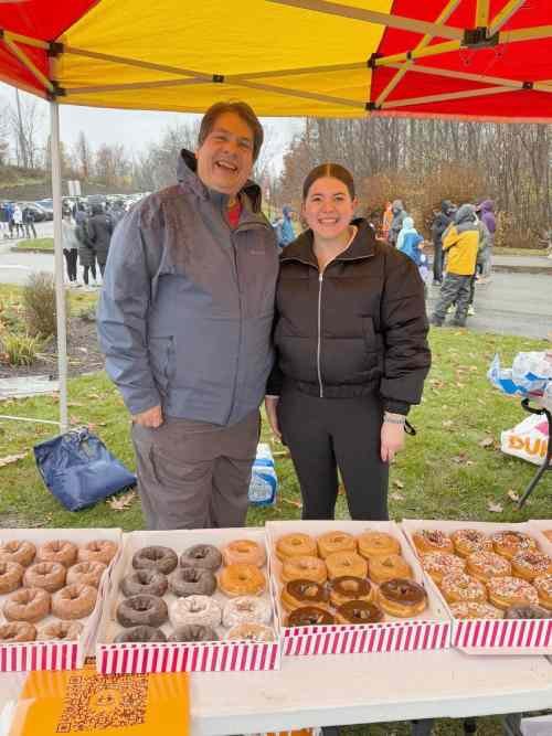 Two smiling people stand behind a table of assorted donuts under a yellow and red canopy at an outdoor event, with people in jackets and trees in the background.