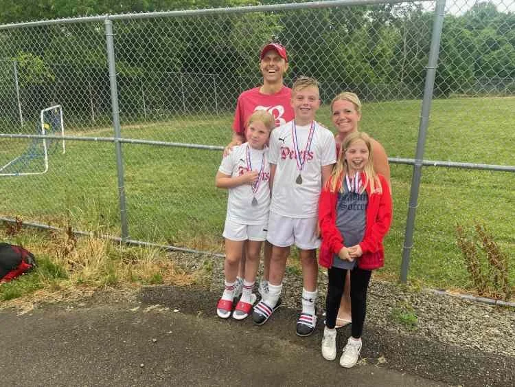 A group of four children and one adult standing outdoors in front of a chain-link fence, celebrating after a sports event. They are smiling, with some wearing medals around their necks. The scene appears to be at a sports field or park.