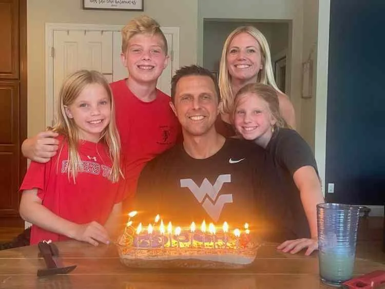 A family of five celebrating a birthday with a cake and lit candles in a home dining area.