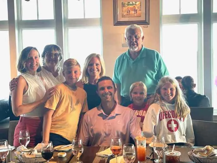 Group of nine people, including children and adults, gathered around a dining table in a restaurant with large windows in the background.