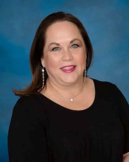 Portrait of a woman with shoulder-length brown hair, wearing a black top and dangling earrings, standing against a blue background.