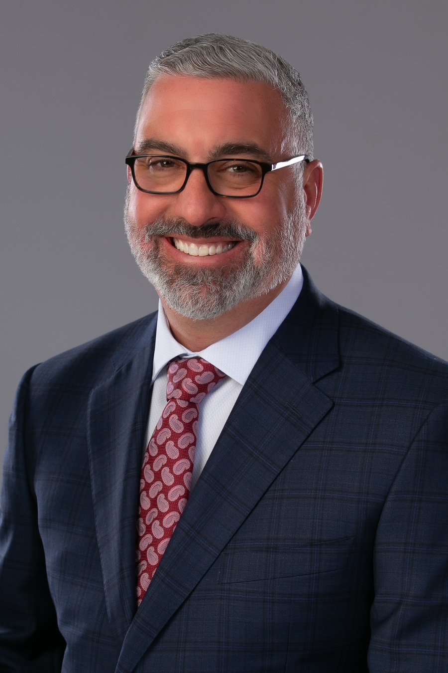 A smiling man with gray hair, beard, and glasses wearing a dark plaid suit, white shirt, and a patterned red tie against a gray background.