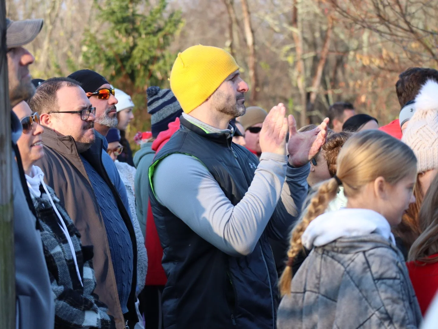 A group of people outdoors during the daytime, some wearing jackets and hats, with various expressions of attentiveness; one man wearing a yellow beanie and clapping.