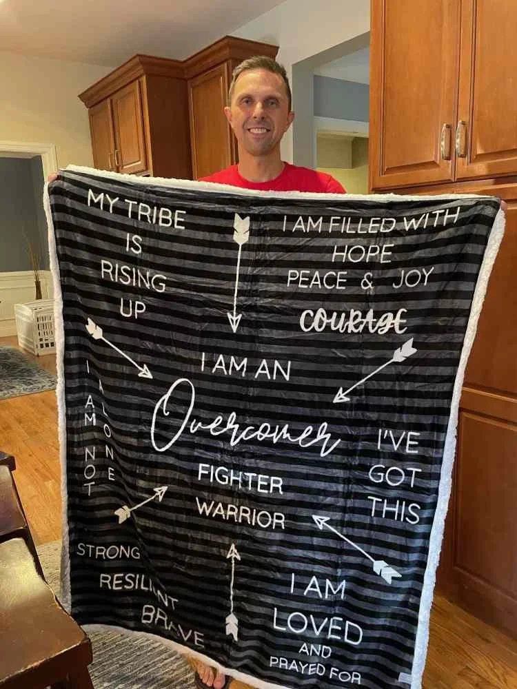 A man standing in a kitchen holding a black and white striped quilt with inspirational text and arrows. The quilt reads, 'My tribe is rising up, I am filled with hope peace & joy courage, I am an acknowledgment, I've got this, I am loved and prayed for, fighter, warrior, strong, resilient, brave.'