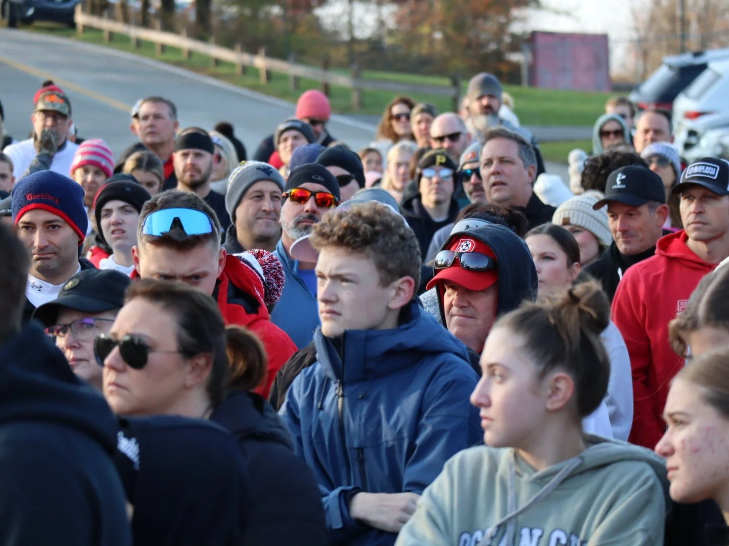 A large group of people gathered outdoors, some wearing jackets, hats, and sunglasses, standing on or near a roadside with cars and trees in the background.