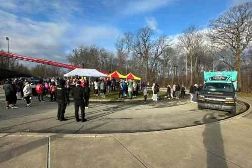 A group of people gathered outdoors around tents and booths on a sunny day with leafless trees in the background and a vehicle parked nearby.