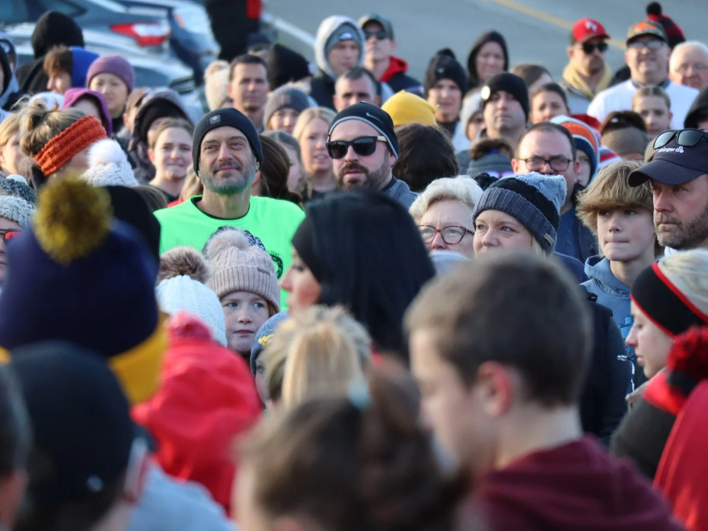 Crowd of people gathered outdoors, wearing winter clothing and hats, some looking forward and others looking around.