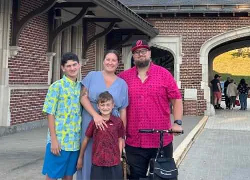 Four people posing outside a brick building with an archway, including two children and two adults, one man holding a bicycle.