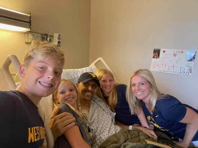 A group of five people, including a young girl in a hospital bed with IV, posing for a selfie in a hospital room.