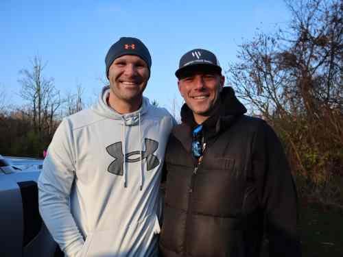 Two men smiling outdoors with clear blue sky, bare trees, and parked cars in the background.