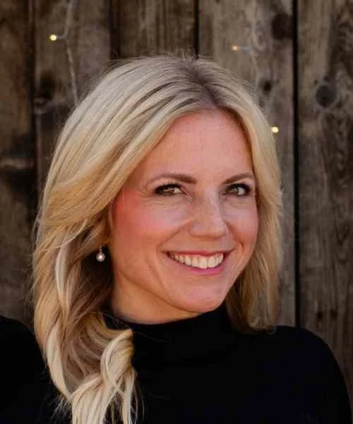 Portrait of a smiling woman with blonde hair, wearing a black top and pearl earrings, standing in front of a wooden fence with string lights.