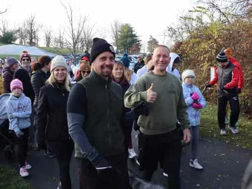 Group of people gathered outdoors, some smiling and giving thumbs up, dressed for cool weather.