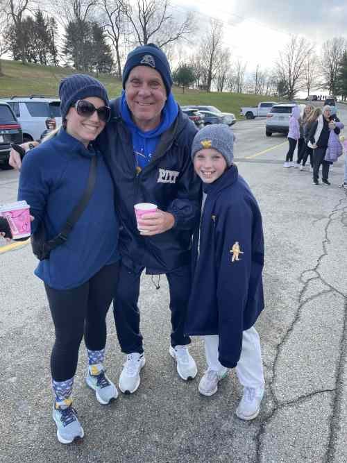 A group of three people, two kids and an adult, smiling at an outdoor event. They are dressed warmly in jackets, hats, and sunglasses, standing on a paved area with parked cars and other people in the background.
