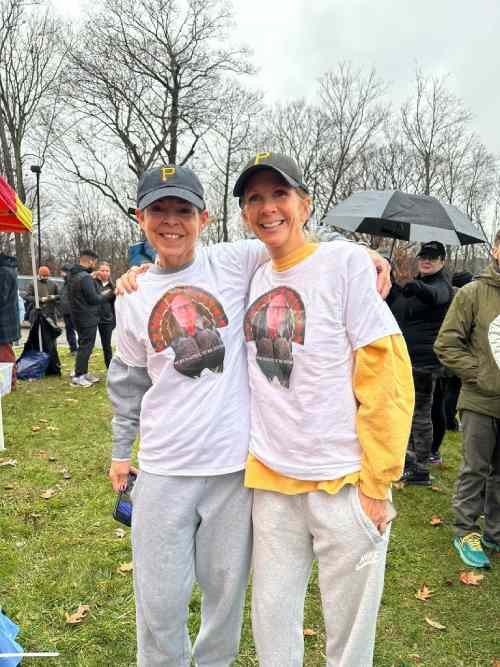 Two women in matching white t-shirts with a graphic image are smiling and standing close together, with their arms around each other, outdoors on a rainy day with leafless trees and other people in the background.