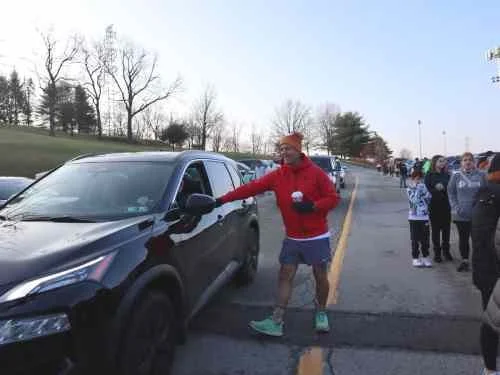 A person in a red jacket and shorts is pointing at a black SUV in a line of parked cars at an outdoor event.