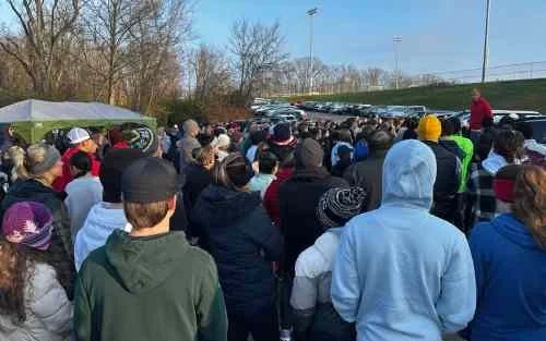 A large crowd of people gathered outdoors, facing a direction out of view, on a clear day with trees and parking lot in the background.