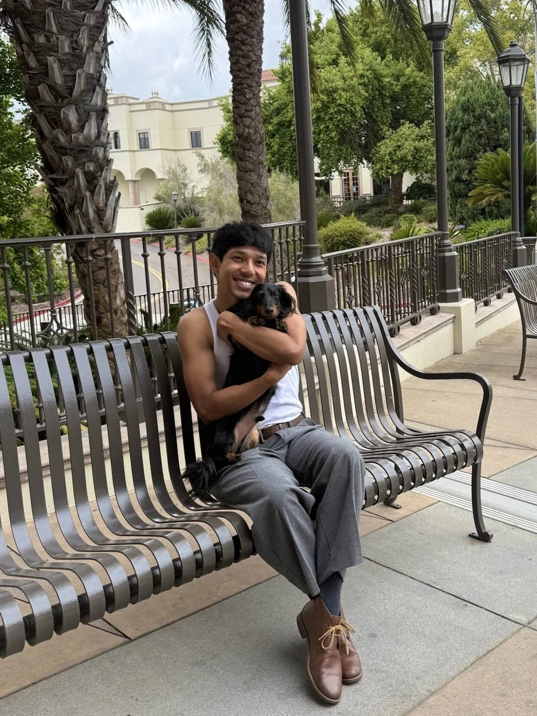 A smiling man in a white tank top and gray pants sitting on a park bench, holding a small black and brown dog. They are outdoors with trees, lampposts, and a white building in the background.