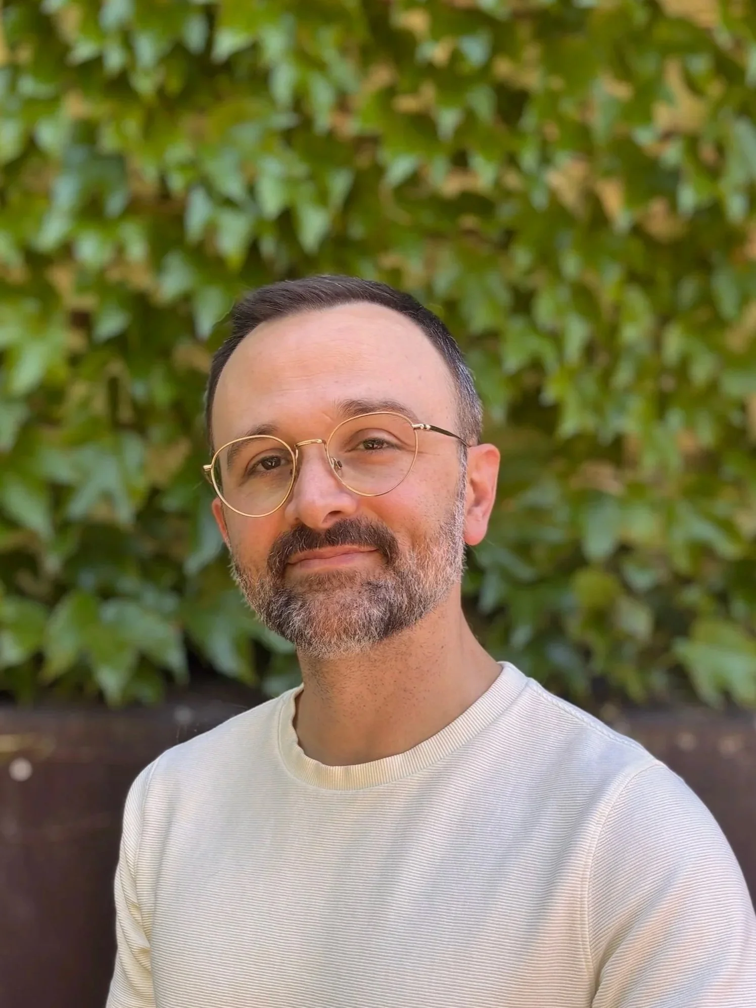 Close-up of a man with glasses and beard, wearing a white shirt, standing outdoors in front of green bushes.