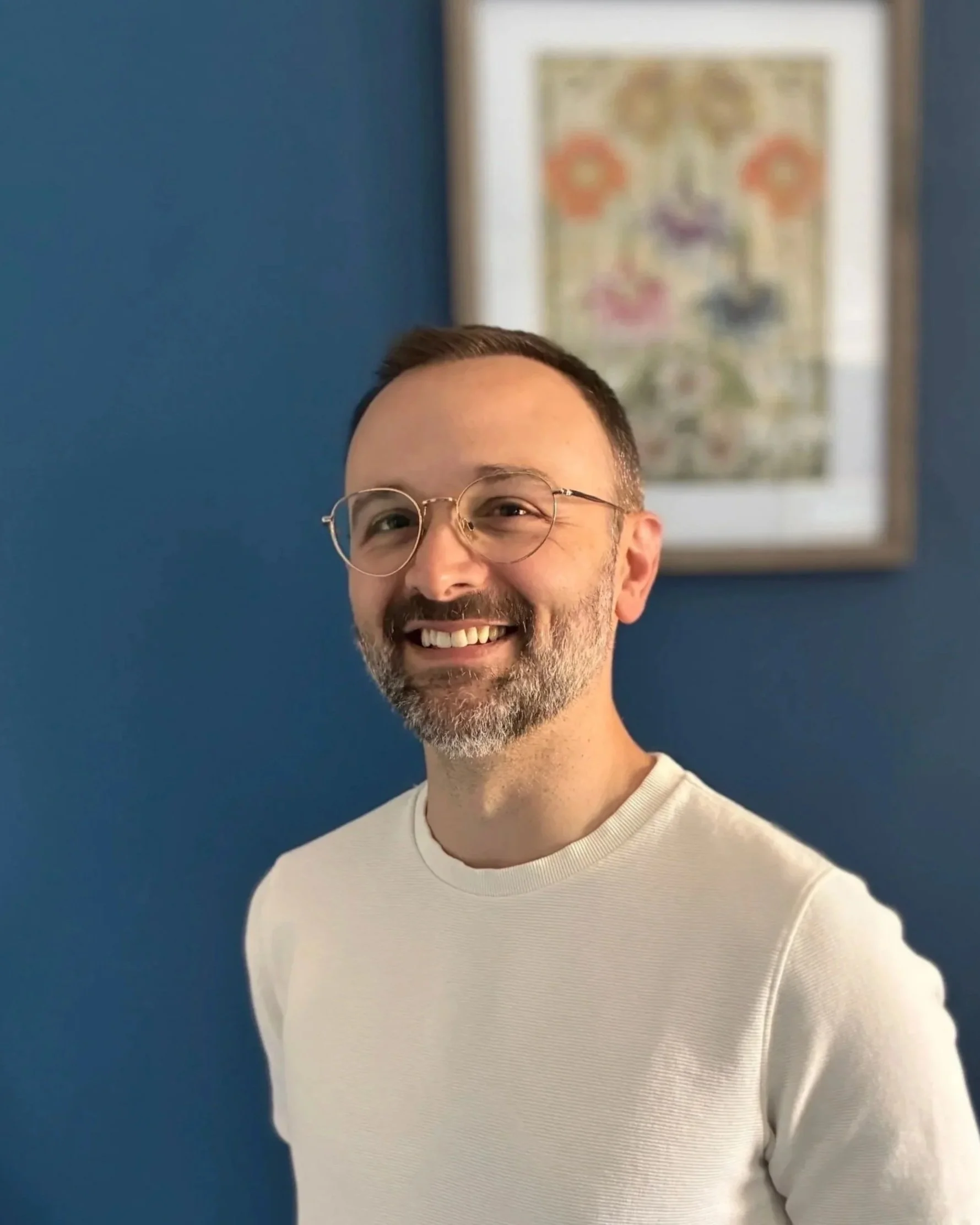 A smiling man with glasses and a beard, wearing a white shirt, standing in front of a blue wall with framed art in the background.