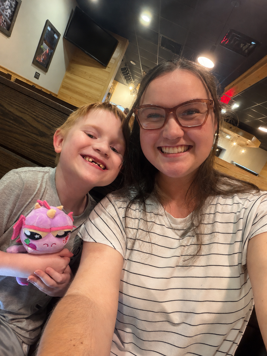 A woman and a young boy smiling for a selfie inside a restaurant or cafe, with a television and wooden decor in the background. The boy is holding a plush toy.