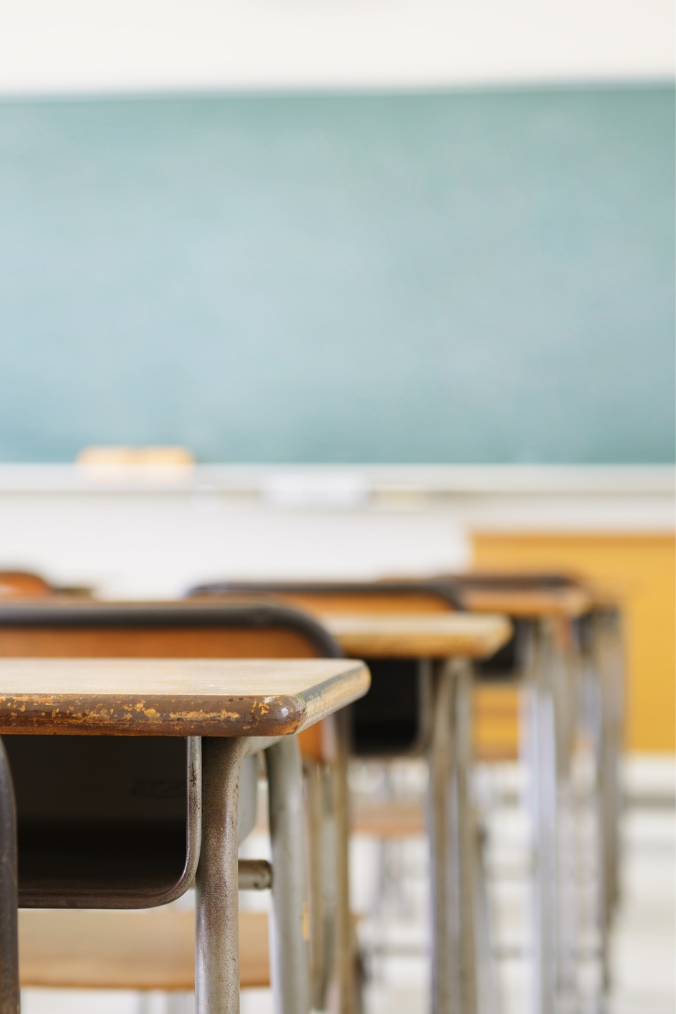 Empty classroom with wooden desks and metal chairs, green chalkboard, and white walls.