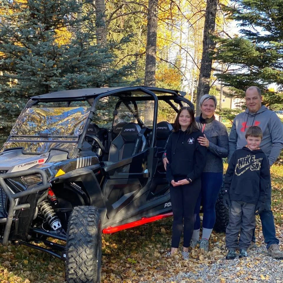 A family of four stands outdoors in a wooded area with fallen leaves, next to a black off-road vehicle, during autumn.