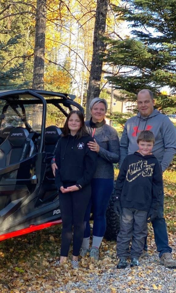 A family of four standing outdoors with trees in the background, next to an all-terrain vehicle. The family includes a woman, a teenage girl, a man, and a young boy, all dressed casually for fall.