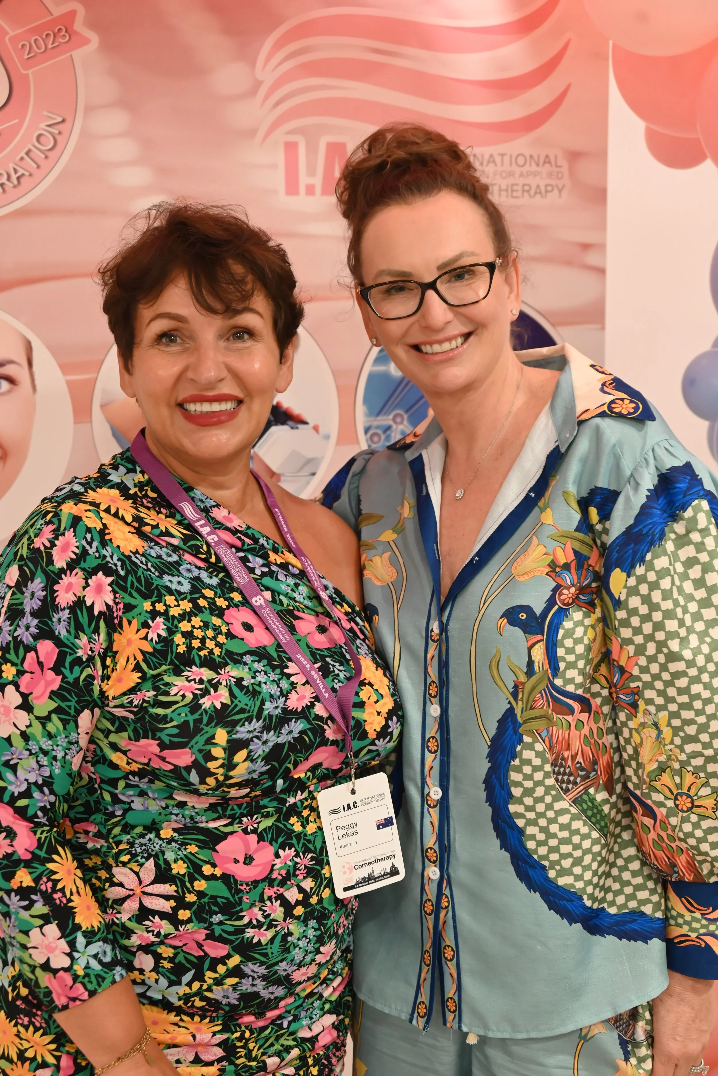 Two smiling women standing in front of a pink backdrop with logos, wearing colorful floral patterns and professional badges around their necks.