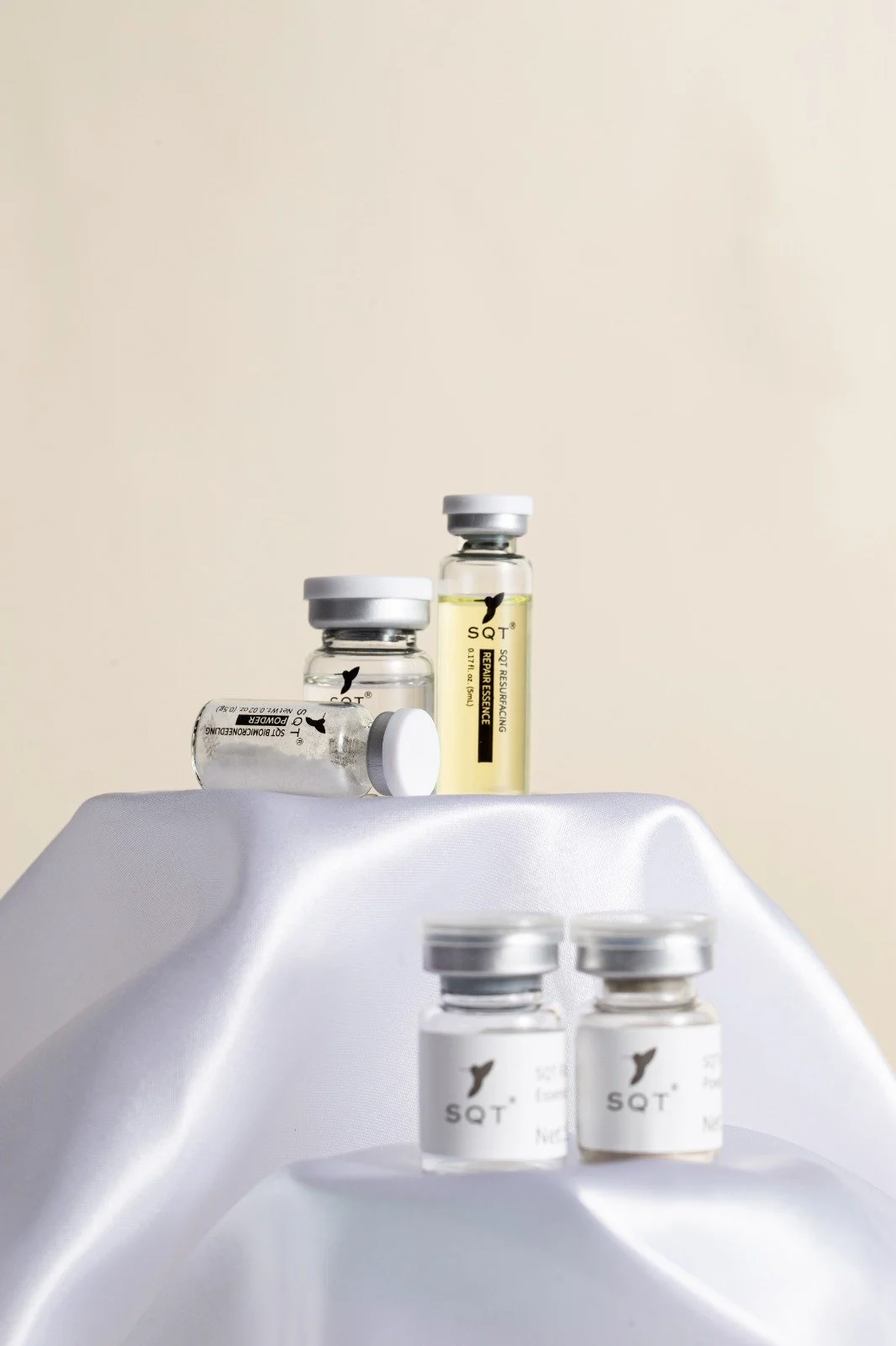 Several small glass bottles with white caps labeled 'SQT' and containing skincare or medication solutions are displayed on a white satin surface against a plain light background.