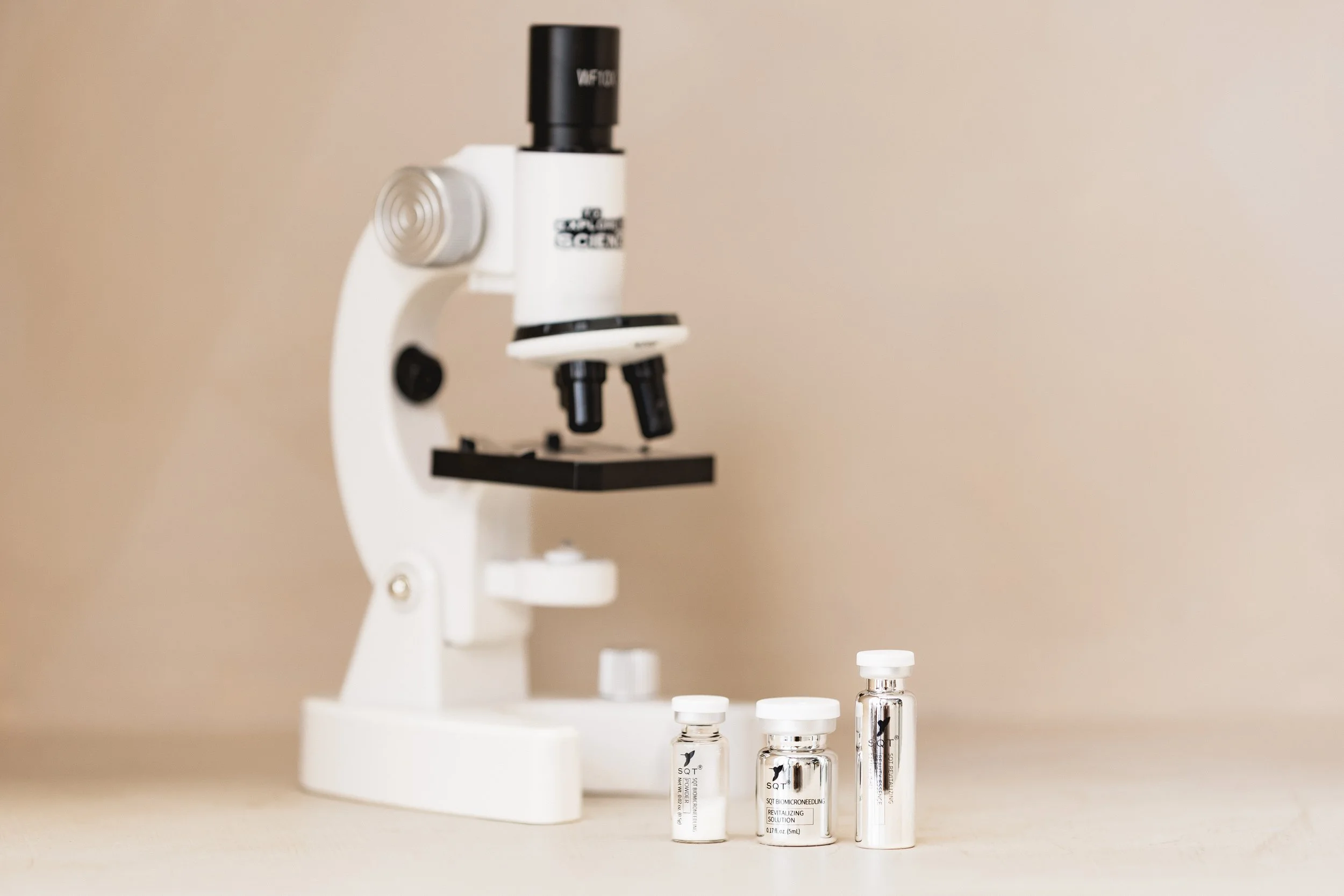 A white microscope with black eyepiece and objective lenses, along with several small bottles of skincare solutions labeled SQY, displayed against a beige background.