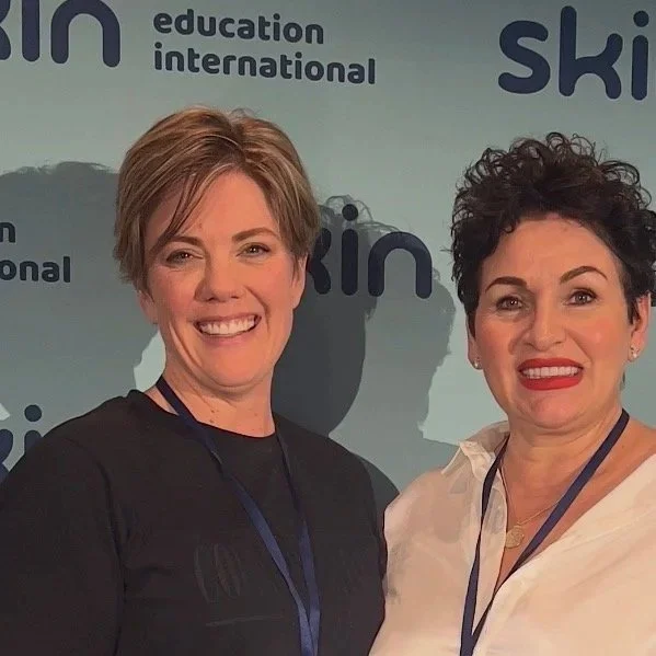 Two women smiling at a conference, wearing name tags, standing in front of a backdrop with the words 'skin', 'education', 'international', and 'ski' visible.