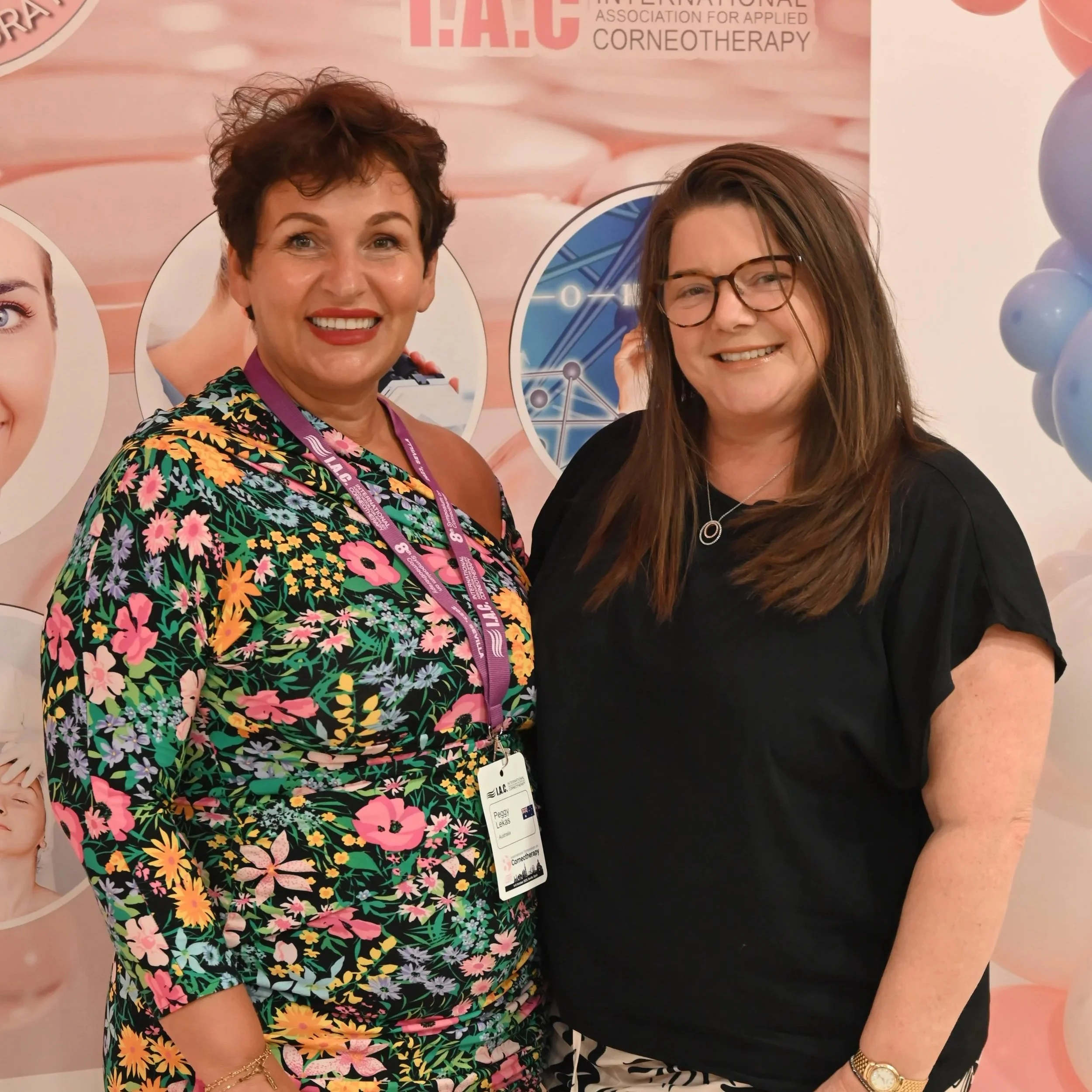 Two women are standing in front of a pink wall that has images of balloons and text related to the International Association for Applied Corneotherapy, smiling at the camera. One woman has short curly hair and is wearing a colorful floral dress. The 