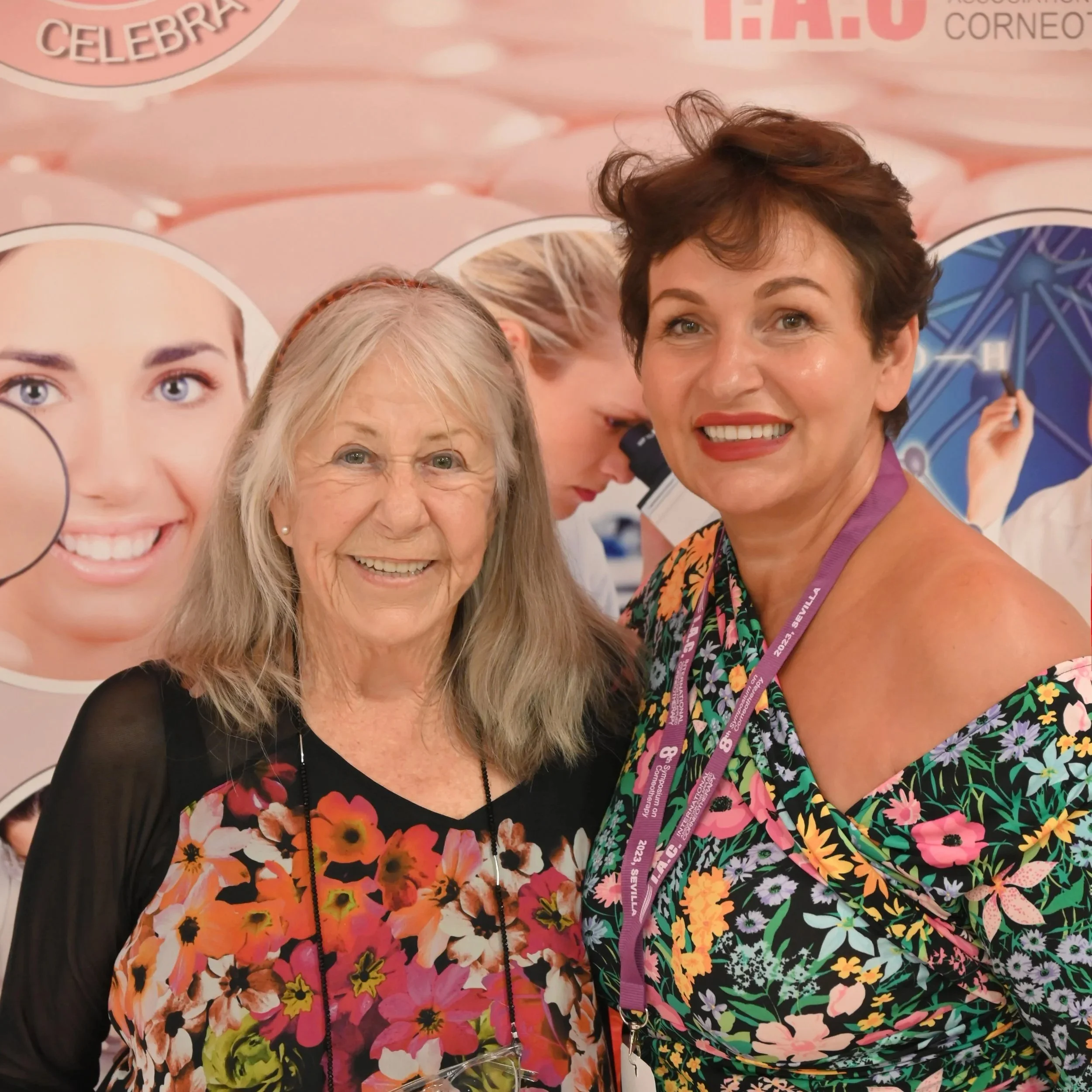 Close-up of three smiling women, one with gray hair and glasses, and two with dark hair, standing in front of a pink background with facial portraits and text, including a lanyard with event details.