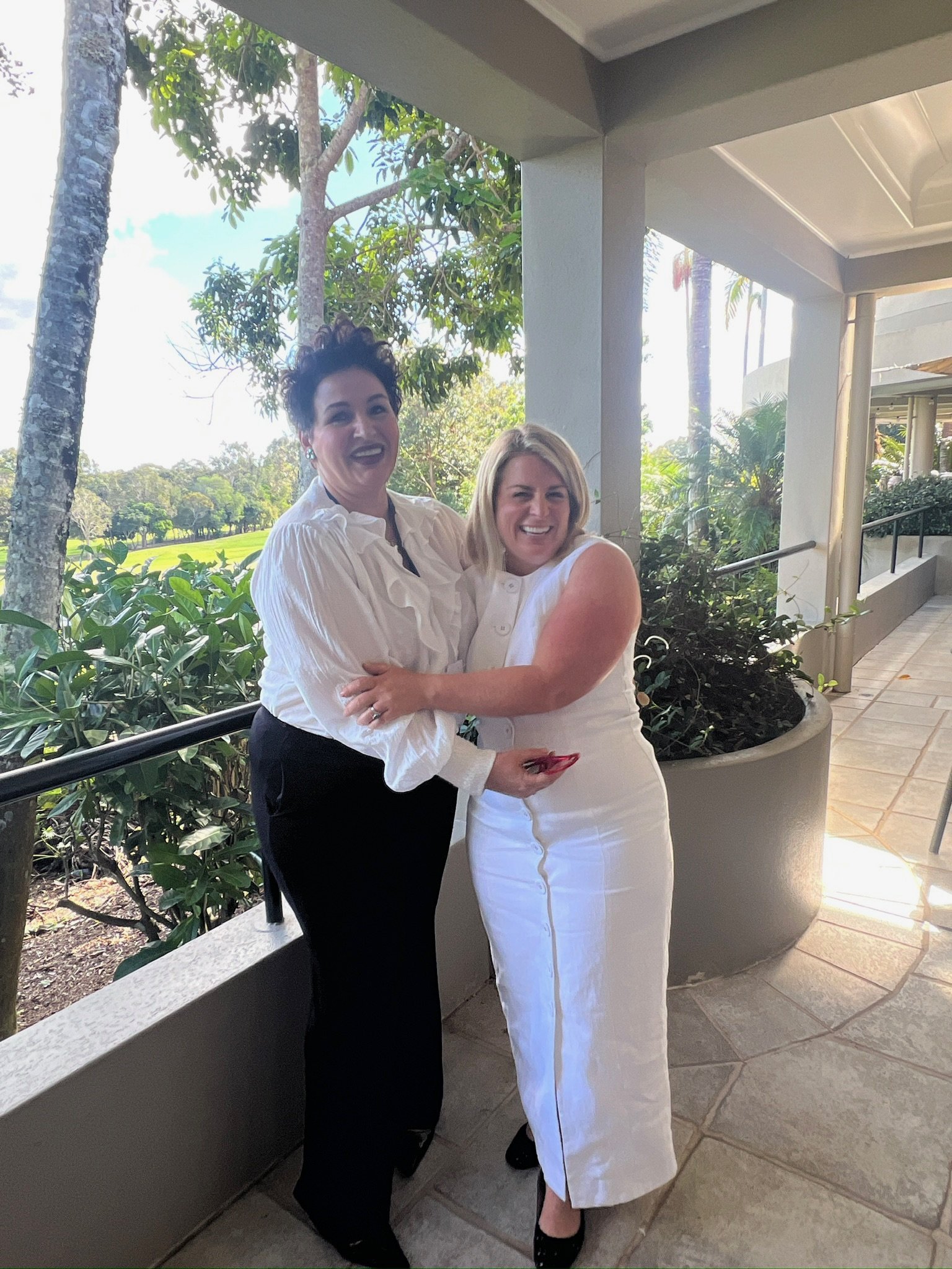 Two women are standing outside on a patio with greenery and trees in the background, smiling and hugging each other.