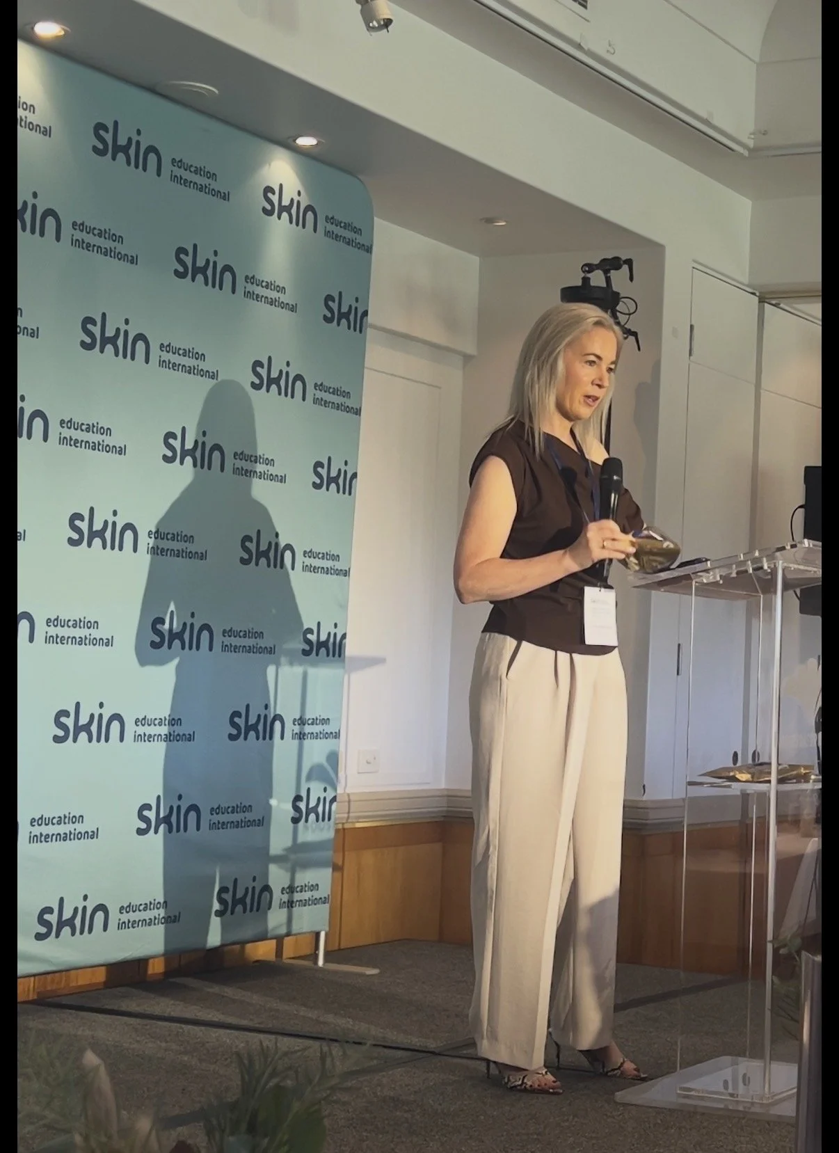 A woman is standing on a stage, holding a microphone and a notebook, giving a presentation at an event organized by Skin International Education. There is a blue backdrop with the repeated logo and the words 'skin' and 'education international'. A sc