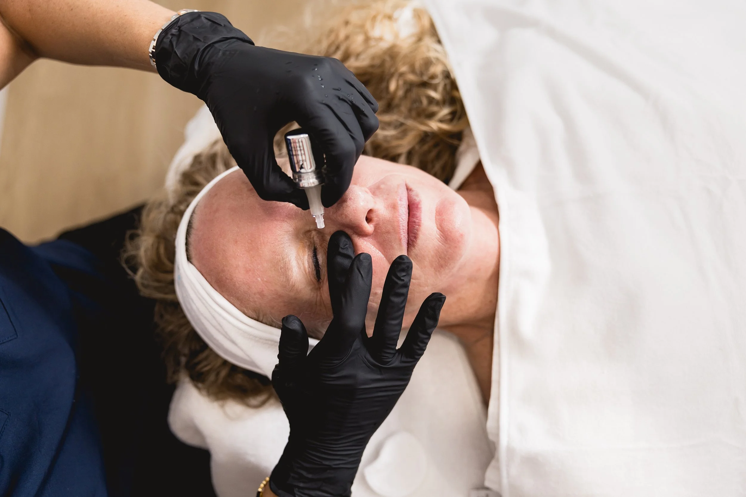 A person receiving a facial treatment while lying down with eyes closed, wearing a headband, and a skincare professional in black gloves applying a serum or treatment to their face with a small dropper.