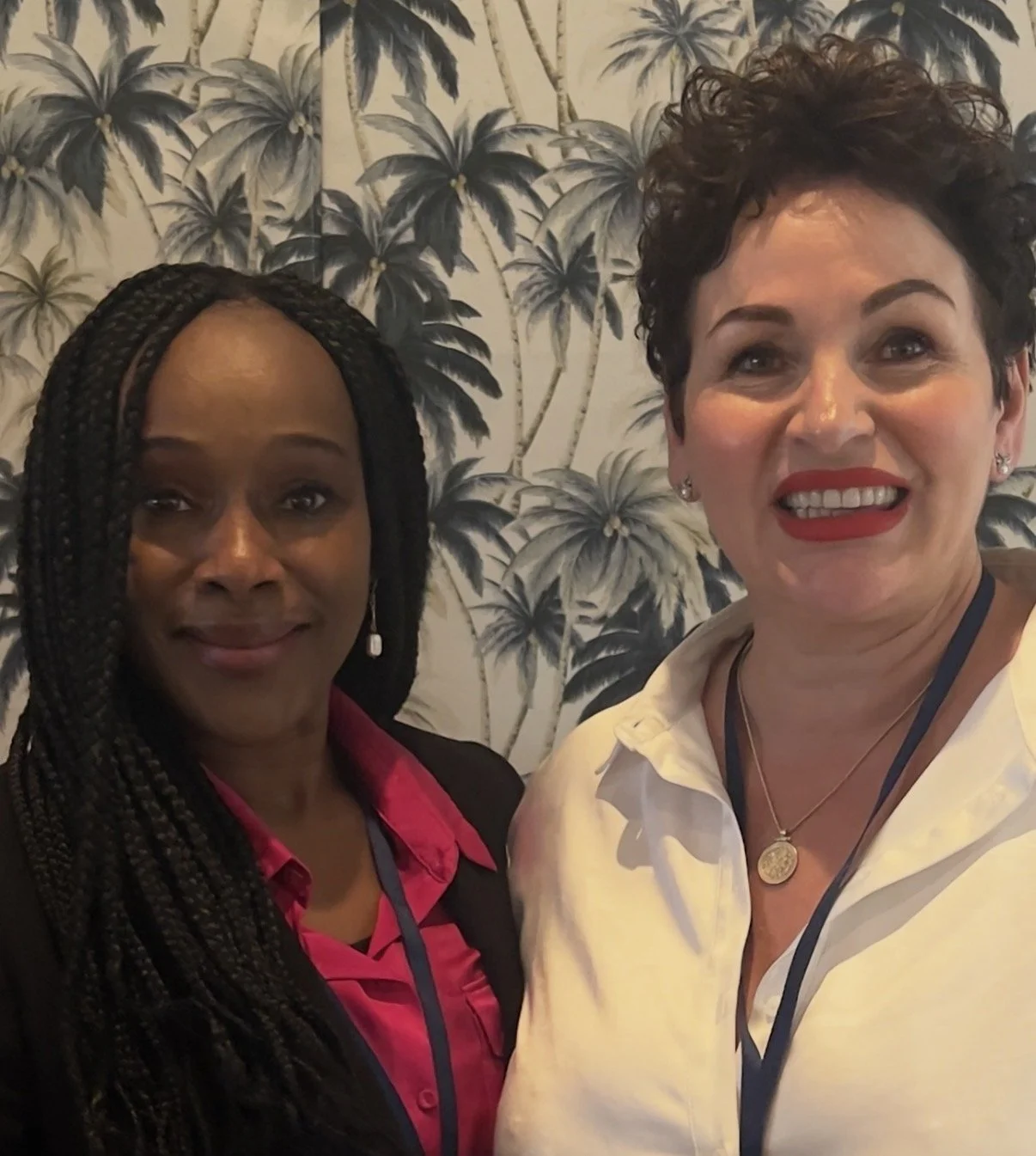 Two women taking a selfie in front of a palm tree patterned wallpaper. One woman has dark skin and braided hair, wearing a black blazer and pink shirt. The other woman has light skin and curly short hair, wearing a white shirt and red lipstick.