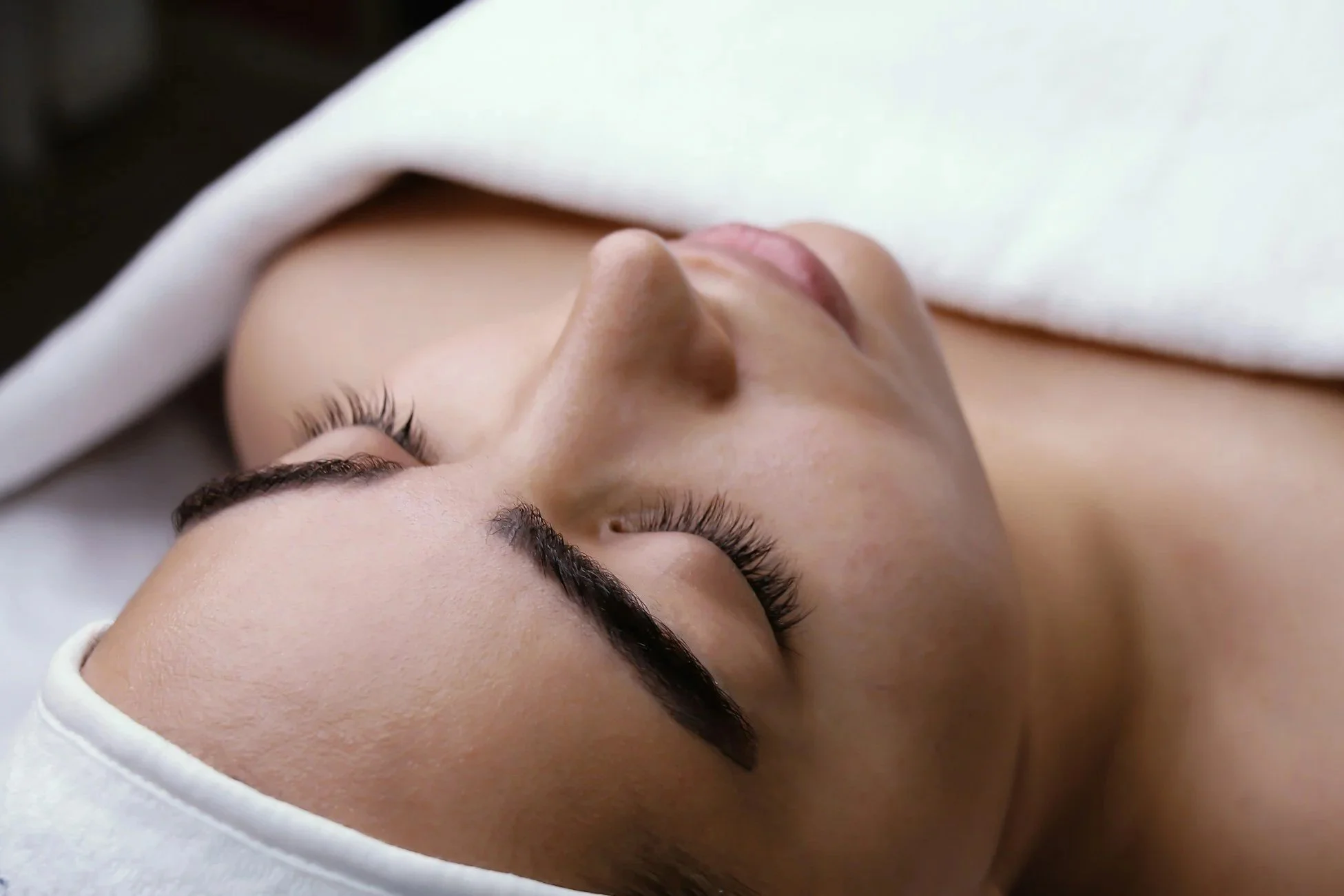 A woman lying down with her eyes closed, receiving a facial treatment. Her skin is smooth, and she is covered with a white towel or cloth around her head.