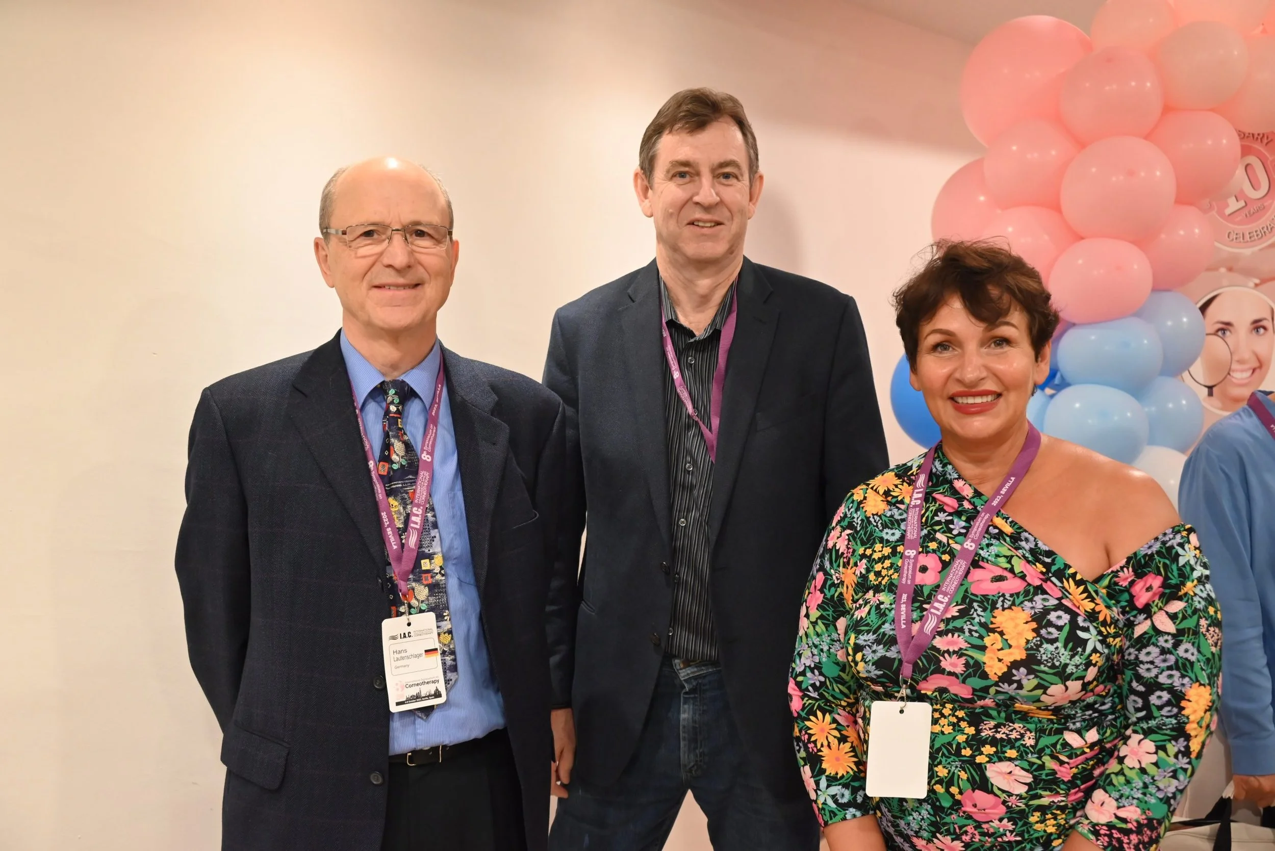 Three people standing together at an event with pink and blue balloons in the background. The man on the left is wearing glasses, a suit, and a colorful tie, with an event badge around his neck. The man in the middle has brown hair and is wearing a d