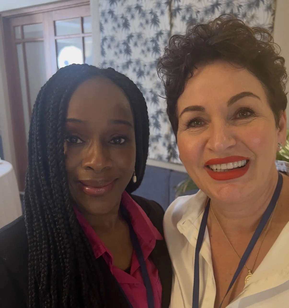 Two women smiling for a selfie indoors, with a window and patterned curtain in the background.