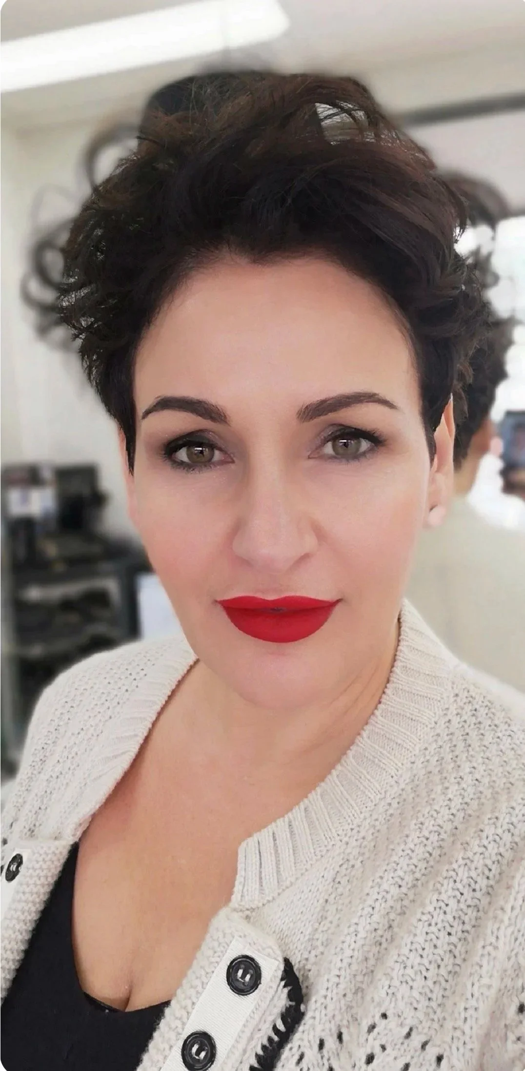A woman with short, dark curly hair and bright red lipstick, wearing a cream-colored cardigan with black buttons, smiling at the camera.