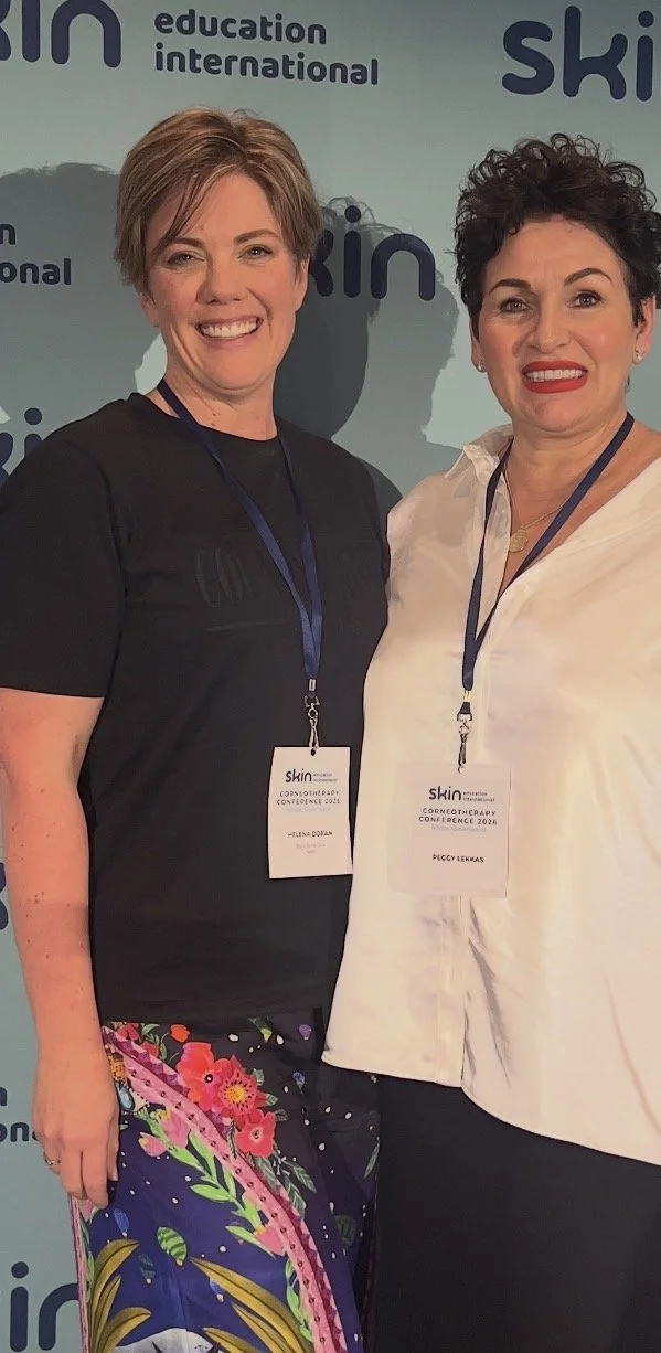 Two women smiling at a conference, wearing name tags, standing in front of a backdrop with the words 'skin', 'education', 'international', and 'ski' visible.