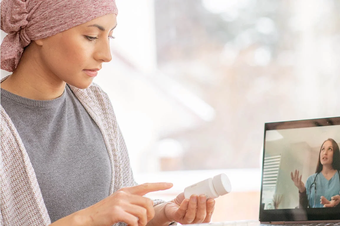 A woman wearing a pink headscarf and grey top is looking at a medicine bottle, while on a video call with a female doctor visible on a laptop screen.