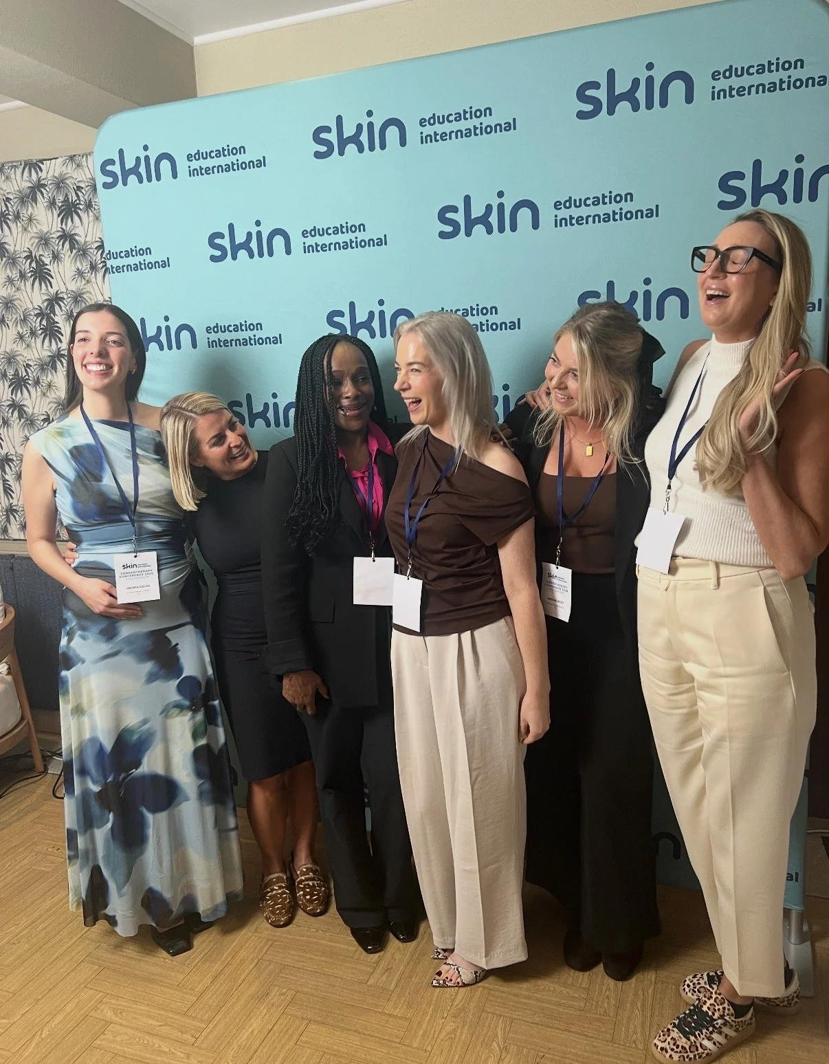 Group of six women standing together in front of a blue backdrop with 'skin education international' printed repeatedly. They are smiling and appear to be at a professional or conference event, wearing business casual attire and name badges.