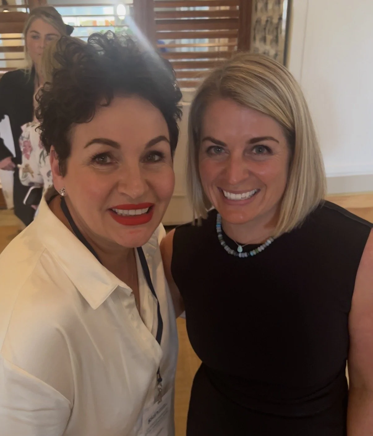 Two women smiling and posing for a photo indoors, with a background of wooden blinds and another woman in the distance.
