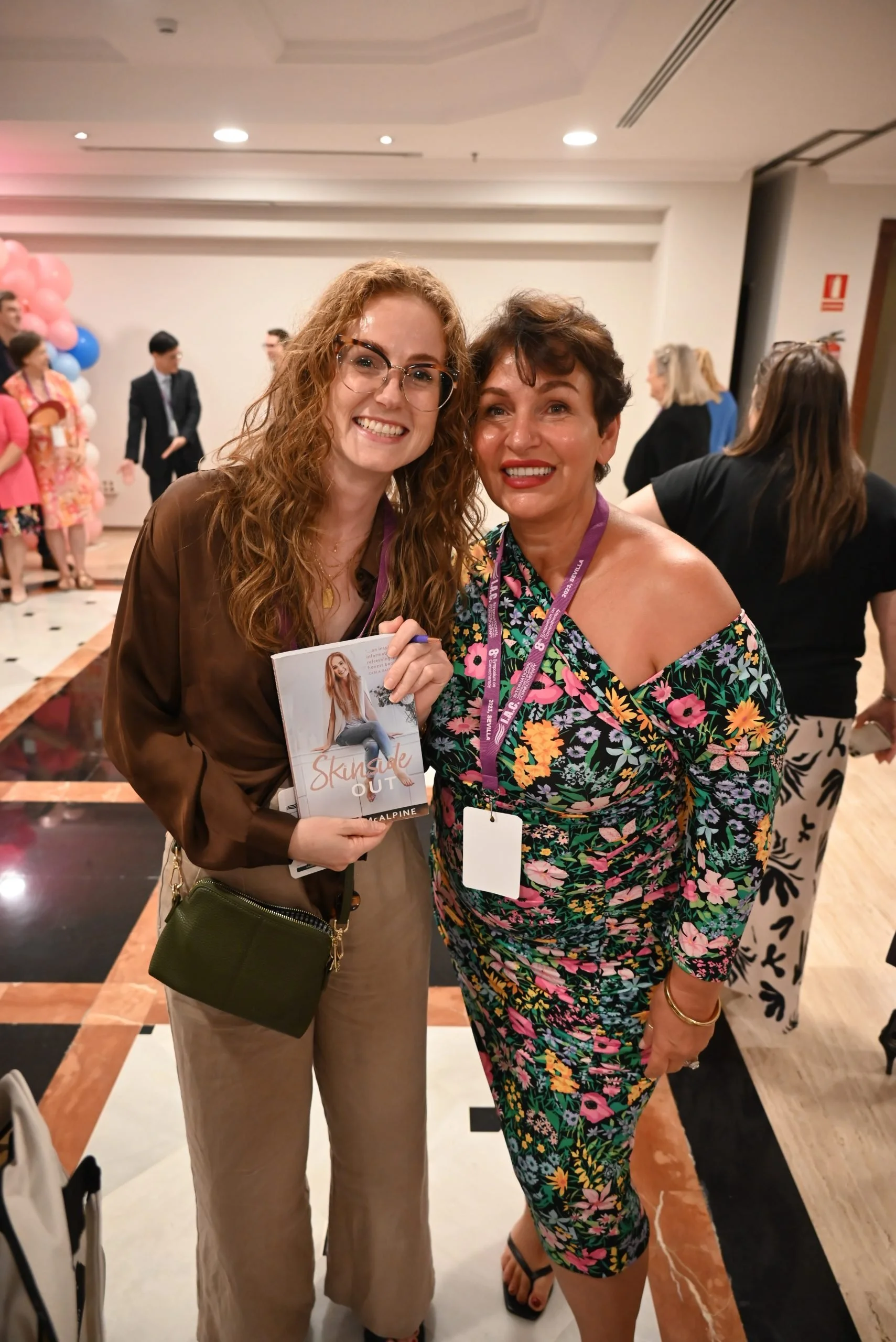 Two women smiling and posing for a photo at an indoor event with other attendees in the background. One woman has curly red hair, glasses, and is wearing a brown blouse and beige pants. She is holding a magazine or booklet. The other woman has short 