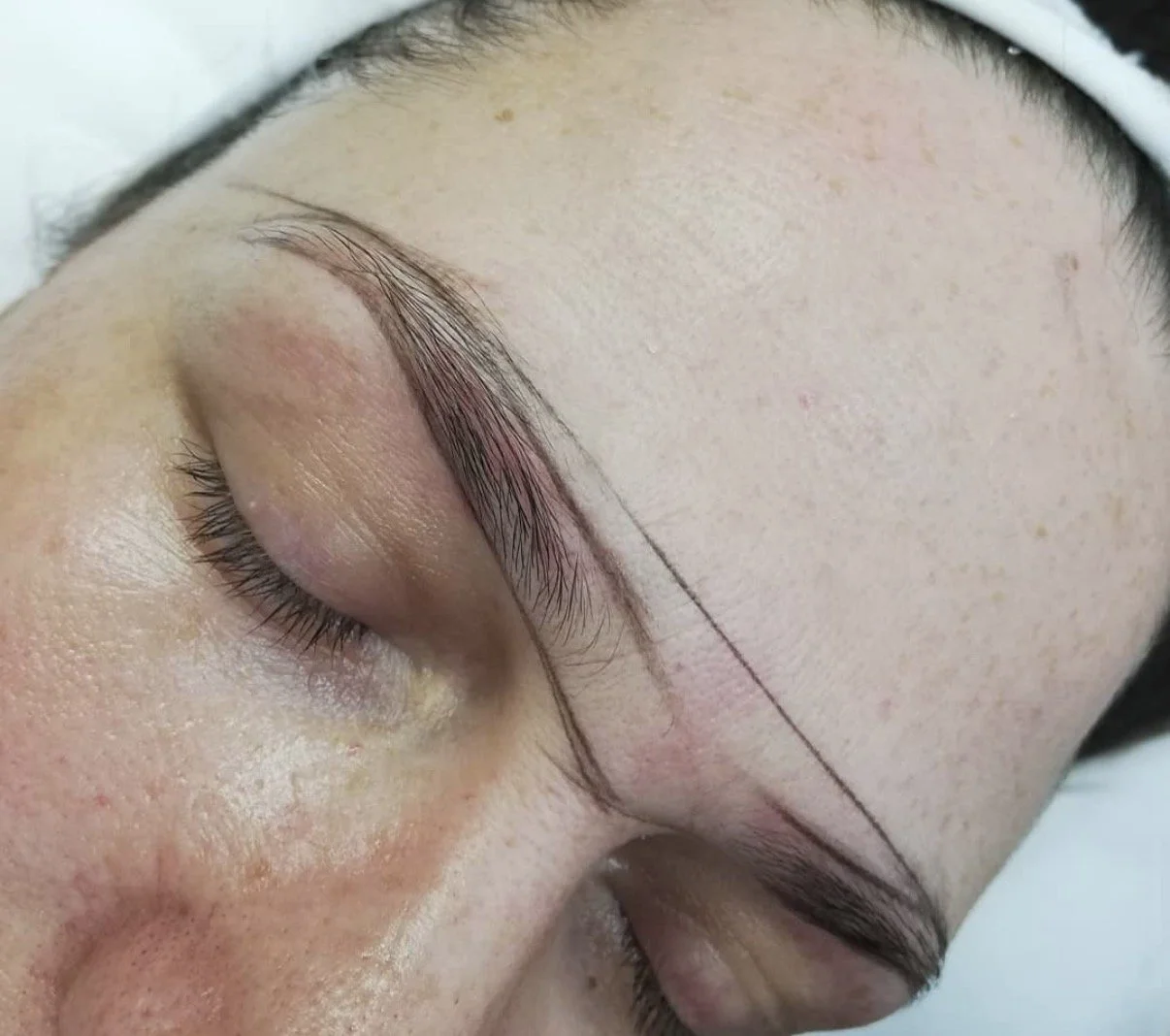 Close-up of a person's face with closed eyes, showing new eyebrow microblading or tattooing.