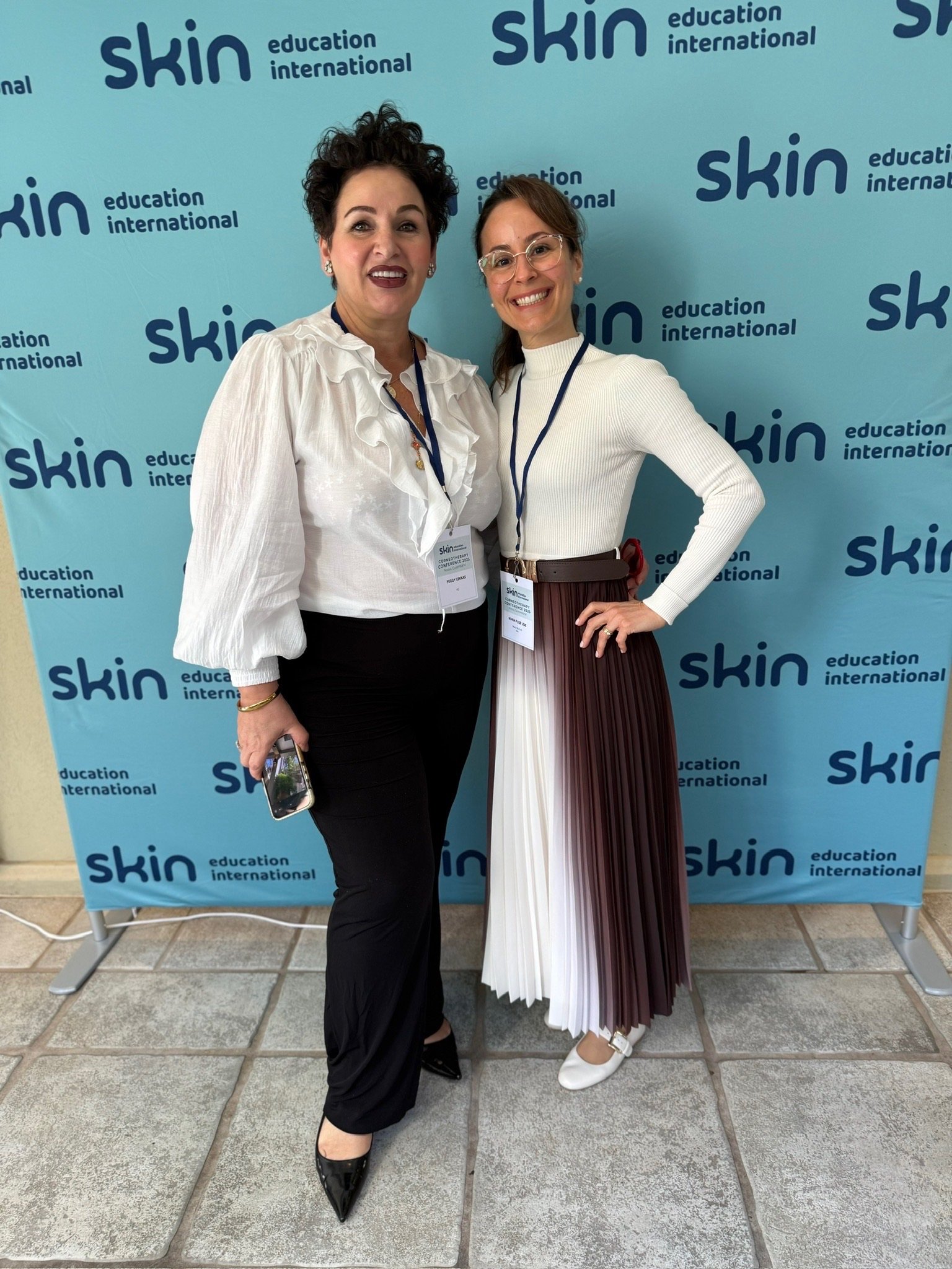 Two women standing in front of a blue backdrop with the words "skin," "education," and "international" repeated. Both women are smiling and wearing glasses, one in a white blouse and black pants, the other in a white turtleneck and pleated multicolor