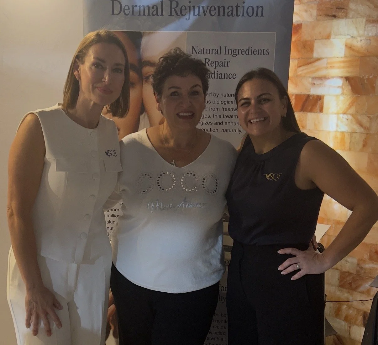 Three women standing together smiling at a skincare event, with a sign behind them that reads "Dermal Rejuvenation" and details about natural ingredients and skin repair.
