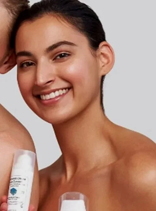 A young woman with dark hair pulled back, smiling, and holding two skincare or cosmetic bottles.
